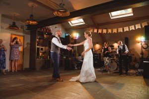 In a warmly lit room adorned for a wedding, the bride in her white gown and the groom in vest and trousers sway to a live band. As guests capture moments, renowned Bristol wedding photographers skillfully frame each enchanting scene.