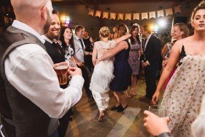 A joyful wedding reception captured beautifully by a documentary wedding photographer, with people dancing and socializing. A woman in a white dress is hugged by another in a blue dress on the dance floor. A Just Married banner hangs in the background, and guests hold drinks, smiling and interacting.