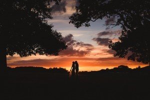 A couple embraces under a vibrant sunset sky, framed by silhouetted trees. Captured by skilled wedding photographers in Somerset, the horizon glows with shades of orange, purple, and pink. The scene conveys a sense of romance and tranquility.
