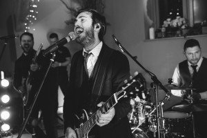 Black and white image of a band performing on stage. The lead singer, playing a guitar, passionately sings into a microphone. Other band members, including a drummer and guitarist, are visible. Captured by talented wedding photographers in Bristol, the setting is festively decorated with lights and plants.