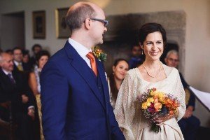 A couple stands at their wedding ceremony, captured beautifully by talented Bristol wedding photographers. The groom wears a blue suit with an orange tie, while the bride is in an ivory dress holding a bouquet of colorful flowers. Guests are seated in the background, smiling and watching.