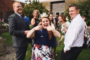Amid the laughter and clinking glasses of a joyful celebration, a woman at the center raises her glass with a radiant smile. The garden setting, framed by rustic buildings, adds to the charm—a scene artfully captured by talented wedding photographers Somerset prides itself on.