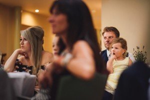 A man in a suit sits next to a young girl in a yellow dress, both watching intently like subjects captured by Somerset wedding photographers. The girl has her finger near her mouth, surrounded by others in a warmly lit setting, possibly a formal event or celebration.