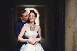 A groom in a navy tuxedo sweetly kisses a smiling brides cheek. The bride, wearing a strapless white gown and large floral necklace, stands in a dimly lit hallway. Captured by talented wedding photographers Somerset, they both appear joyful and affectionate.