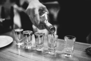 A person is pouring a clear liquid from a bottle into five shot glasses on a wooden surface. The black-and-white image, reminiscent of those captured by wedding photographers in Somerset, exudes a classic, timeless feel.