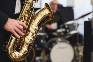 A close-up of a person playing a golden saxophone captures the essence of live music. In the blurred background, another performer is seated at a drum set. The focus on the saxophones shiny details reflects the artistry often sought by Bristol wedding photographers in capturing special moments.