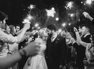 A newlywed couple shares a kiss beneath a canopy of sparklers held by smiling guests. The black-and-white photo, captured by talented wedding photographers from Bristol, perfectly encapsulates the romantic and celebratory atmosphere as friends and family surround the couple in an evening celebration.