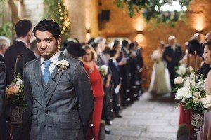 A groom in a gray suit stands at the altar with a thoughtful expression, captured beautifully by skilled Somerset wedding photographers. The aisle is lined with flowers, guests are seated on either side, and in the warmly lit background, a bride in white approaches, accompanied by a man.