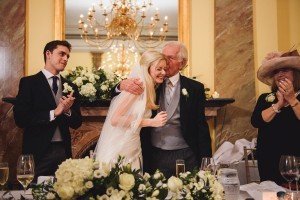 A bride in a white gown and veil is hugged by an elderly man in a suit, both smiling. They are surrounded by three people applauding. Captured beautifully by a documentary wedding photographer, the elegant setting features a chandelier and floral decorations reminiscent of the Cotswolds charm.