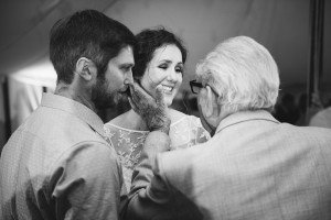 An elderly man gently touches a bearded mans face while a woman in a lace dress smiles at them. Captured by talented documentary wedding photographers, theyre under a tent in this grayscale photo, radiating warmth and connection.