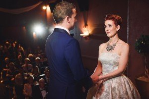 A bride and groom stand facing each other, holding hands. The bride wears a strapless gown and necklace, while the groom wears a blue suit. Theyre in a dimly lit room with onlookers seated behind them. Captured perfectly by Somerset wedding photographers, the lighting highlights the couple beautifully.