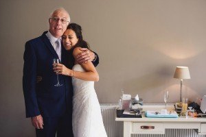 A bride in a white dress smiles and embraces an older man in a suit, whos holding a champagne glass. They stand in a warmly lit room with a cluttered desk and lamp, captured beautifully by documentary wedding photographers.