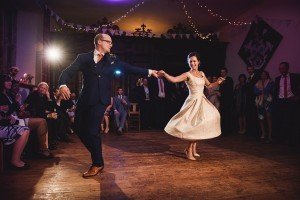 A couple dances joyfully on a wooden floor under string lights at a celebration, elegantly captured by Somerset wedding photographers. The woman wears a light dress, the man is in a suit, and guests watch and smile in the dimly lit room.