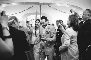 A joyful couple walks down an aisle in a festive celebration, captured beautifully by Bristol wedding photographers. The groom, smiling in a light-colored suit, holds the brides hand as guests cheer and toss confetti. The bride, beaming in her white gown amid the lively crowd inside a decorated venue, radiates happiness.