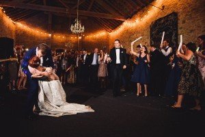 A couple shares a romantic dance in the center of a rustic venue, with guests in formal attire watching and celebrating. String lights and a chandelier illuminate the scene, creating a warm, festive atmosphere captured beautifully by wedding photographers from Bristol.