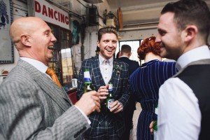 A group of three men, dressed in formal suits, are happily chatting and holding beer bottles at an indoor gathering. One man, in a checkered suit, is smiling broadly with a sign that reads DANCING visible in the background, capturing moments like skilled Somerset wedding photographers.