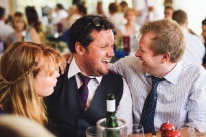 Three people are sitting closely at a gathering, smiling and laughing joyfully, as if captured by skilled documentary wedding photographers. Two men are wearing dress shirts and ties; one also has a vest and sunglasses atop his head. A woman with light brown hair sits next to the men. A wine bottle is on the table.