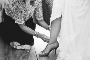 A woman in a lace top kneels on the floor, helping another adjust her high-heeled shoe. The second woman, dressed in a white gown with lace details, showcases an intimate moment captured beautifully by Somerset wedding photographers. The black-and-white image exudes timeless elegance.