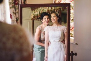 Two women smiling in front of a mirror. One wears a white wedding dress with lace details and a tiara, while the other is in a light green dress with embroidery. Floral curtains are visible in the background—a perfect moment captured by Somerset wedding photographers.