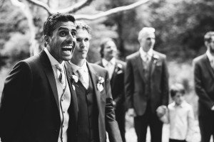 A grayscale image of a wedding scene outdoors captured by documentary wedding photographers. A groom, smiling broadly, stands beside another man in a suit. Three more men in suits and a child holding a ball are visible in the blurred background, surrounded by trees.