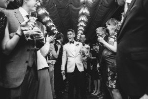A groom in a white jacket walks joyfully through a crowd of well-dressed guests holding drinks. Captured by renowned wedding photographers from Somerset, everyone celebrates under a tent, creating a festive, elegant atmosphere. The images black-and-white palette enhances its timeless charm.