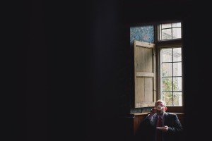 A person in a suit sips from a cup while sitting by a window, the wooden shutters open to let in soft natural light. The dimly lit room offers a serene ambiance, perfect for capturing the moment—a favorite spot for Somerset wedding photographers seeking candid elegance with greenery outside.