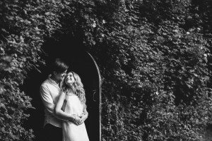 A couple embraces while standing in a doorway framed by lush greenery. The woman, wearing a light dress, smiles at the man behind her with his arms around her waist. Captured in black and white by talented wedding photographers in Bristol, the setting is serene and intimate.
