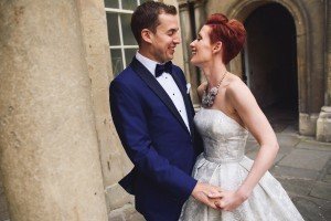 A couple, dressed elegantly, smiles at each other while holding hands in a stone courtyard—a moment beautifully captured by documentary wedding photographers. The man dons a blue suit with a bow tie, and the woman is in a strapless silver gown with a statement necklace.