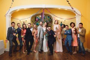 A joyful wedding group poses indoors, captured artfully by a talented documentary wedding photographer. The couple stands at the center, surrounded by friends and family. Everyone is smiling and celebrating, with colorful streamers and decorations in the background against a yellow wall.