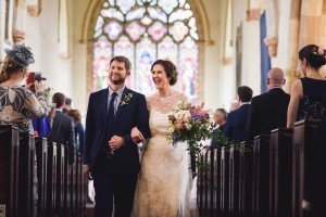 A bride and groom joyfully walk down the aisle of a church with stained glass windows behind them, captured beautifully by wedding photographers Bristol cherishes. The bride holds a bouquet of flowers as guests, some snapping photos, celebrate this special moment. The atmosphere is truly magical.