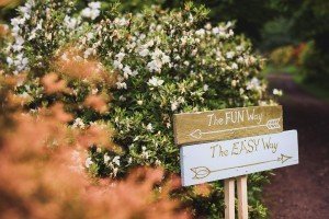 Two wooden signs with arrows point the way amidst white flowers. The top reads The FUN Way, and the bottom says The EASY Way. Green foliage surrounds them, and a dirt path lies beyond—a picturesque scene perfect for Bristol wedding photographers to capture.