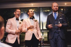 Three men stand together at an event, captured by a talented Somerset wedding photographer. Two men in pink tuxedos laugh and hold champagne glasses, while the man in a gray suit appears to be giving a speech, holding a piece of paper and looking amused.