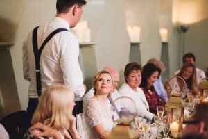 A groom in suspenders stands giving a speech at a wedding reception while the bride, seated, looks up at him smiling. Guests are seated around a long table adorned with candles and lanterns, as Somerset wedding photographers capture the heartfelt moments everyone is listening attentively to.
