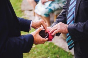 In a picturesque outdoor setting, two individuals in suits gracefully exchange a ring. One hand cradles a small box while the other gently places the ring on a red cushion. Captured through the lens of talented Somerset wedding photographers, a person in a floral dress watches from the background.