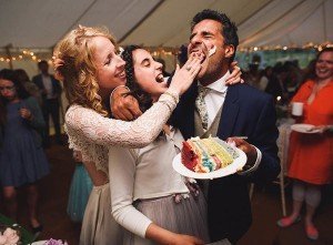 In a joyful celebration scene captured by renowned Bristol wedding photographers, a man in a suit is playfully fed colorful cake by smiling friends inside a beautifully decorated tent. Meanwhile, guests in the background revel in the festivities, cherishing memories that will last a lifetime.