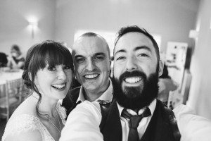 A black-and-white photo of three people smiling for a selfie. A bearded man, like a documentary wedding photographer, holds the camera, joined by a woman with bangs and a man in a suit, inside a room with softly lit walls and chairs in the background.