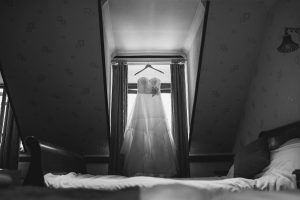 A wedding dress hangs elegantly on a hanger in front of a window, framed by curtains, in an attic room with sloped ceilings. The dim lighting casts shadows across the bed in the foreground. Captured by a talented Bristol wedding photographer, this black-and-white photo exudes timeless charm.