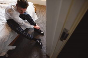A person in a white shirt and plaid pants sits on a bed tying black shoes, perhaps prepping for a days work as one of the wedding photographers Bristol relies on. The room, with its carpeted floor and white walls, has an unmade bed partially visible in the backdrop.