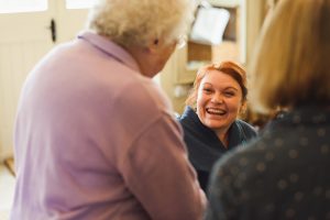 A woman with red hair laughs, facing an elderly person with white hair in a purple sweater, reminiscent of intimate moments captured by documentary wedding photographers. Another person with brown hair is partially visible, their indoor setting complete near a door.