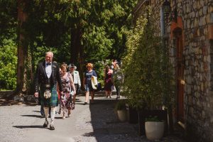A group of people dressed in formal attire, possibly captured by wedding photographers from Bristol, walk along a gravel path next to a stone building. The lead figure wears a kilt. Trees and bushes line the path, with sunlight filtering through the foliage.