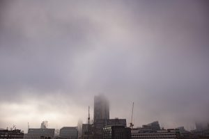 Foggy cityscape with tall buildings partially obscured by mist. In the foreground, a variety of structures stand while cranes suggest ongoing construction. The moody, overcast sky offers an atmospheric backdrop perfect for documentary wedding photographers capturing unique urban moments.
