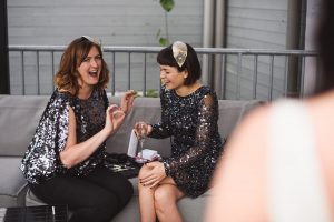 Two women sit on a sofa, laughing together in sequined tops and headbands, as if theyre models posing for Somerset wedding photographers. Makeup items are scattered around, hinting at preparations. A metal railing and light gray wall frame their joyful moment.