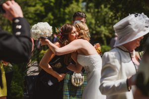 A bride in a white dress embraces a woman in a black dress holding a bouquet of white flowers. They are outdoors, surrounded by greenery and wedding guests, while documentary wedding photographers capture the moment. Nearby, a man in a suit and a woman in a white hat smile warmly.