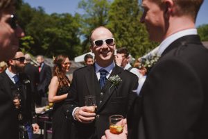 Amidst the lush greenery and clear skies of the garden party, a lively group in formal attire shares stories. In the center, a person with sunglasses and a drink is joyfully engaging with others, perfectly captured by top documentary wedding photographers from Bristol.