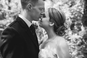 In this timeless black and white photo by Somerset wedding photographers, a bride in a strapless dress with a birdcage veil and her groom in a suit share an intimate moment outdoors, their noses touching amid blurred foliage.