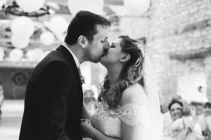 A bride and groom kiss at their wedding, captured by talented Bristol wedding photographers. The black and white image showcases the couple in formal attire, with a veil-clad bride in a lace dress. Guests blur into the warmly lit venue with stone walls, creating an intimate, timeless scene.