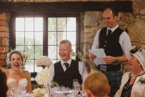A joyful wedding scene unfolds as the bride and groom laugh at a table. A man in a kilt stands, giving a speech, while another woman smiles in her hat. This rustic room with stone walls and a window captures the perfect moment for any documentary wedding photographer in the Cotswolds.