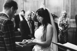 In a wood-paneled room, captured flawlessly by wedding photographers Somerset, a bride and groom exchange rings. The bride, graced with a veil and tiara, stands under the watchful eyes of guests. This timeless black-and-white image embodies the essence of their special day.