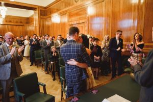 A couple dressed for a formal occasion embraces passionately in a wood-paneled room, surrounded by clapping guests. The scene is celebratory, captured by talented wedding photographers from Somerset, with attendees smiling, suggesting its a wedding or similar event.