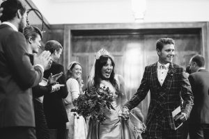 A joyful couple, dressed elegantly, walks down the aisle in a stylish indoor venue captured beautifully by wedding photographers from Bristol. The bride holds a bouquet and wears a tiara, while the groom dons a checked suit. Guests, some clapping, are standing and smiling in the background. Black and white photo.